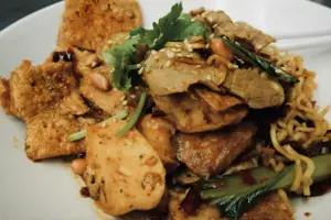This dish features a hearty mix of noodles, thin slices of meat, and fried tofu garnished with fresh cilantro and sesame seeds. It's tossed with peanuts and bok choy in a savory, chili-flecked sauce for a vibrant variety of textures and flavors.