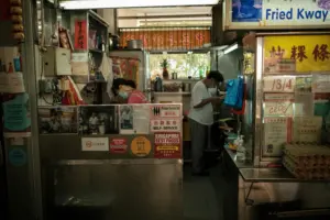 This photograph captures the narrow, bustling interior of a Singaporean hawker stall, where two workers in face masks are busy preparing food behind a stainless steel counter. The space is densely packed with kitchen equipment, egg cartons, and numerous accolades and signs, including one prominently advertising "Fried Kway Teow."