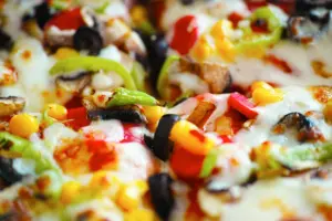 This close-up shot captures a vibrant pizza topped with melted white cheese and a colorful array of fresh vegetables. Visible ingredients include sliced black olives, golden corn kernels, green bell peppers, and savory mushrooms.