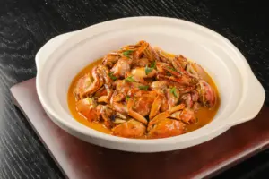 This image features a clay pot filled with crab segments simmered in a rich, orange-hued savory sauce. Small green herbs are scattered over the dish, which is presented on a dark wooden board against a textured black background.