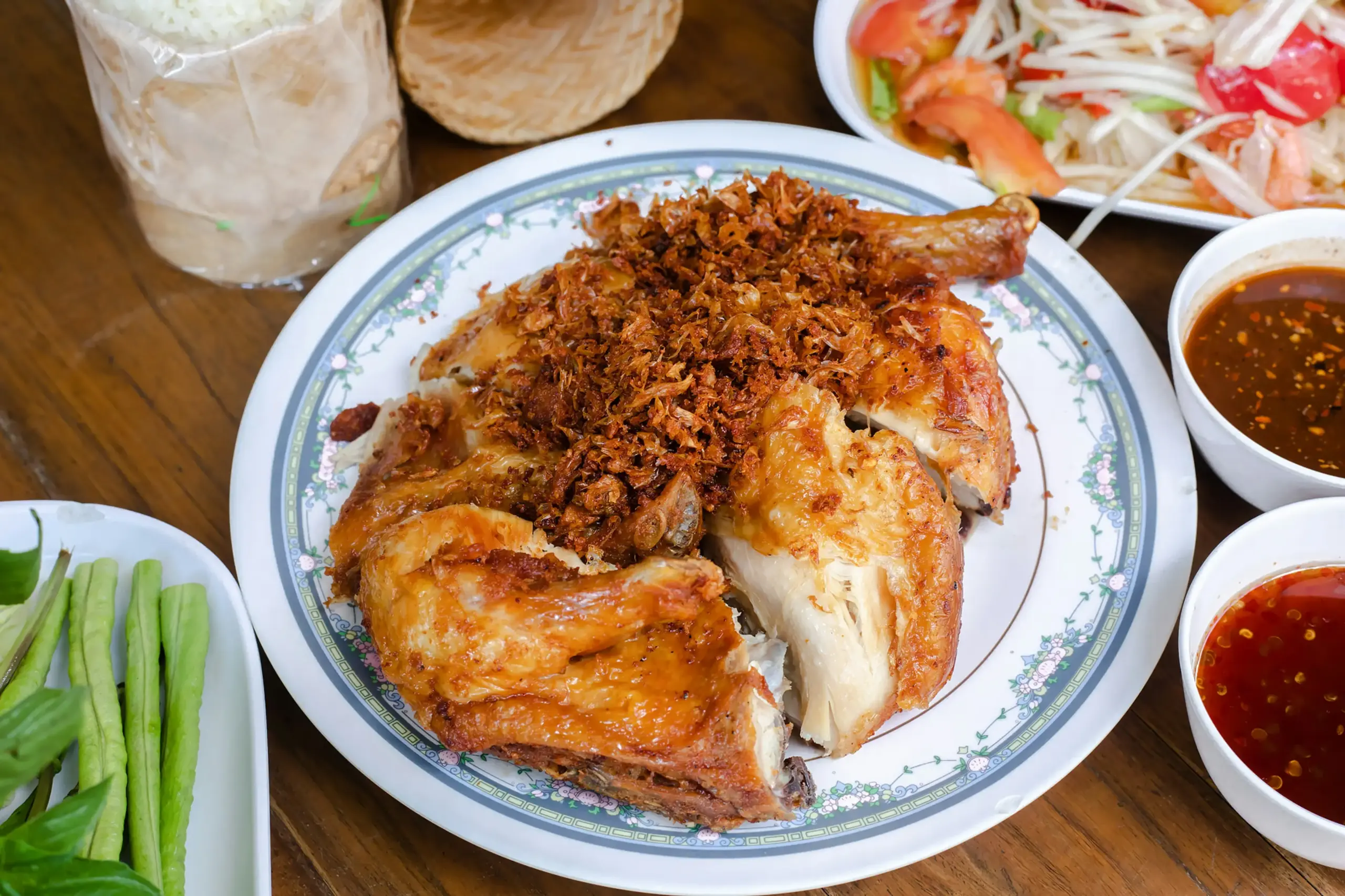 A plate of golden-brown fried chicken topped with crispy garlic sits at the center of a traditional Thai meal. Accompanying the dish are sides of papaya salad, sticky rice in a woven basket, fresh long beans, and two varieties of dipping sauce.