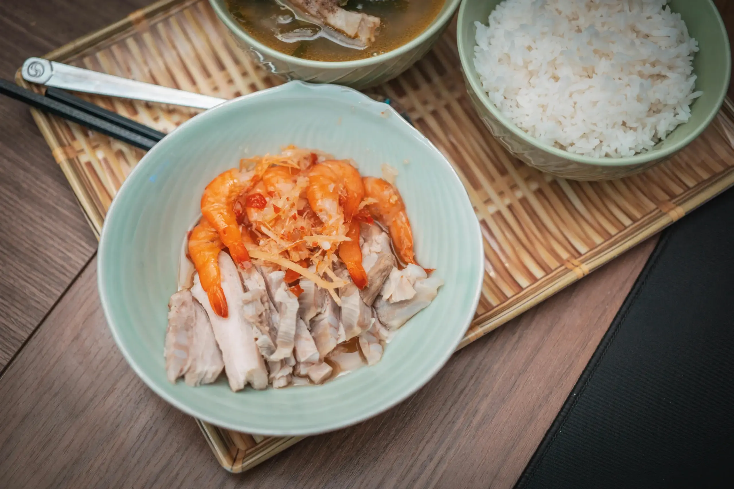 This high-angle shot shows a meal featuring a light green bowl of sliced pork and whole shrimp topped with shredded ginger and chili. Accompanying the main dish is a bowl of white rice and a savory soup, all arranged on a woven tray with a pair of chopsticks.