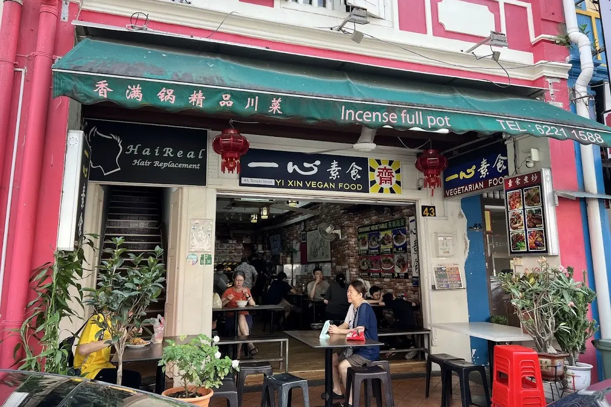 This image captures the exterior of Yi Xin Vegan Food, a humble restaurant located at 43 Temple Street with an open-front seating area. Patrons are seen dining at simple tables under a green awning, flanked by a hair replacement shop and vibrant pink building walls.
