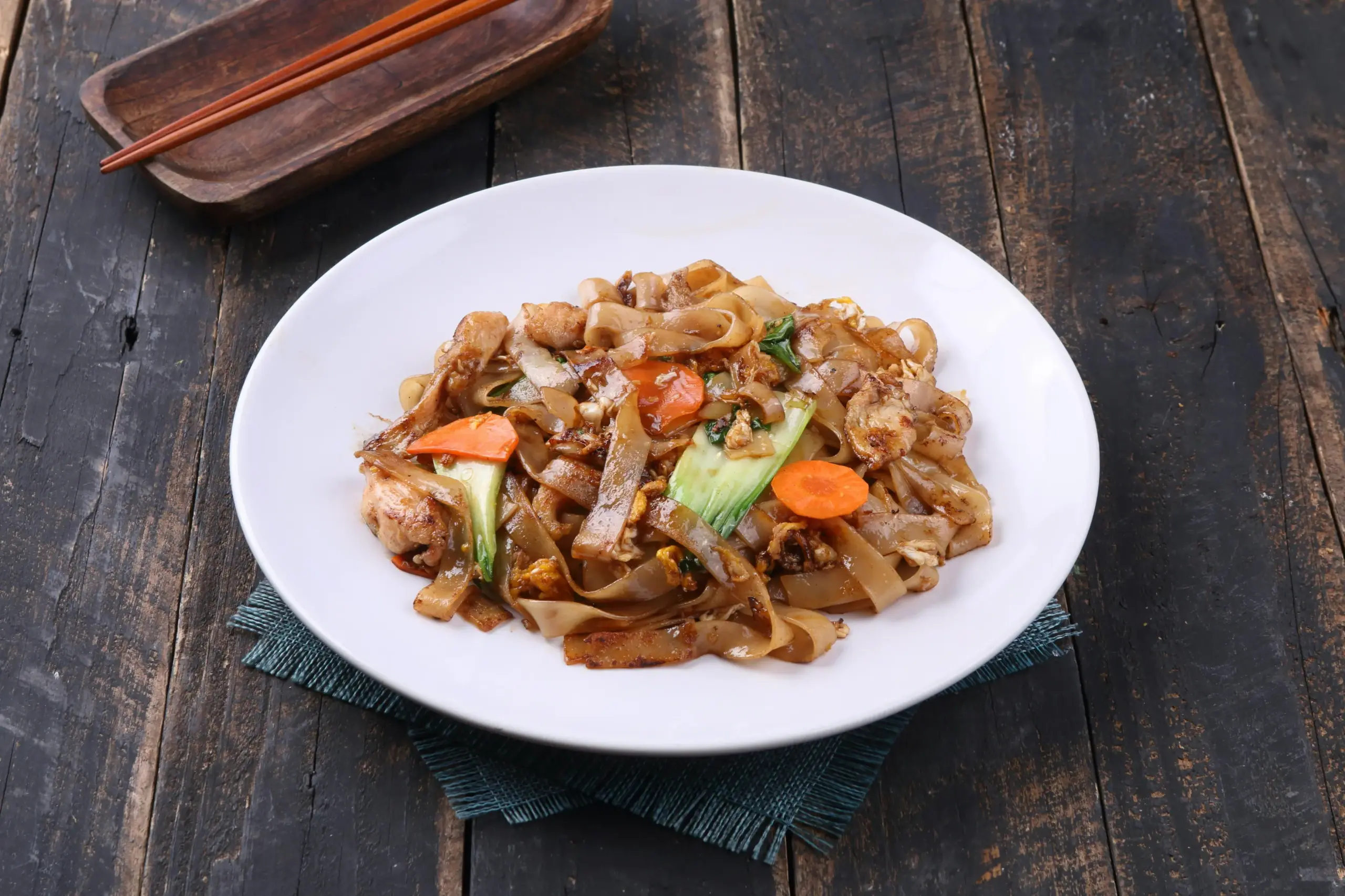 A plate of stir-fried wide rice noodles is mixed with sliced carrots, leafy greens, and protein in a savory sauce. The dish is served on a white plate atop a dark wooden table, accompanied by a pair of wooden chopsticks resting on a side tray.