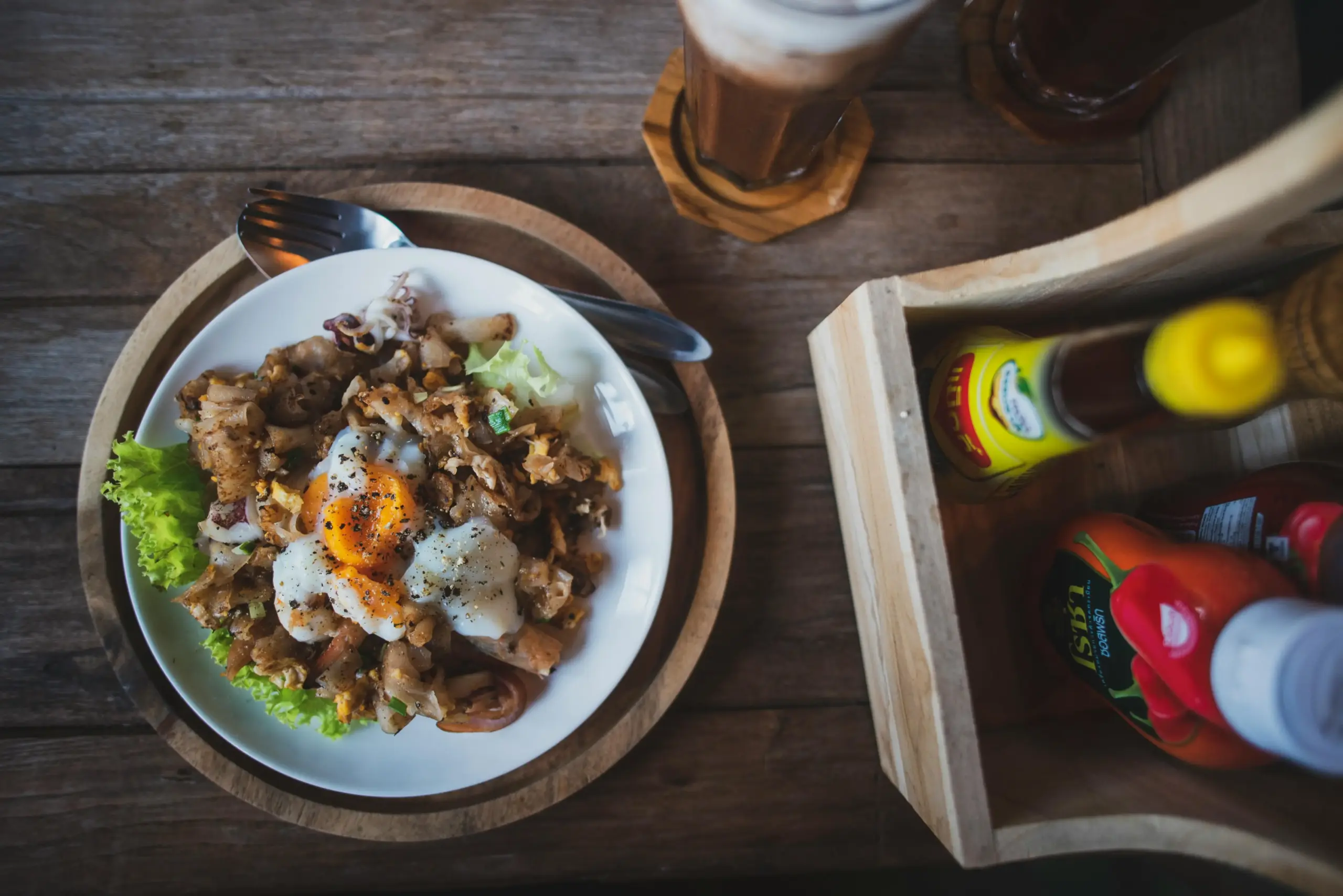 A plate of stir-fried flat rice noodles is topped with a soft-cooked egg and served on a rustic wooden table alongside a tall iced drink. To the right, a wooden condiment caddy holds bottles of chili sauce and other seasonings, completing the casual dining scene.