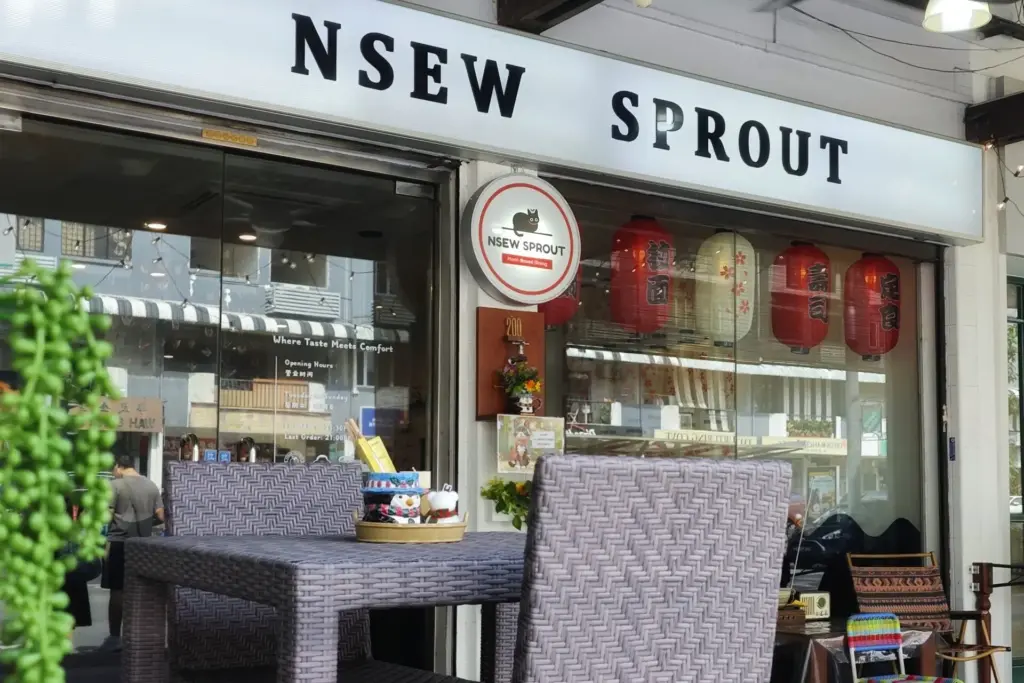 The image shows the exterior of a plant-based restaurant named NSEW Sprout, featuring a prominent white sign and a circular logo with a black cat. Outdoor seating with grey wicker chairs and a decorated table is positioned in front of the glass storefront, which is adorned with red and white Japanese-style lanterns.