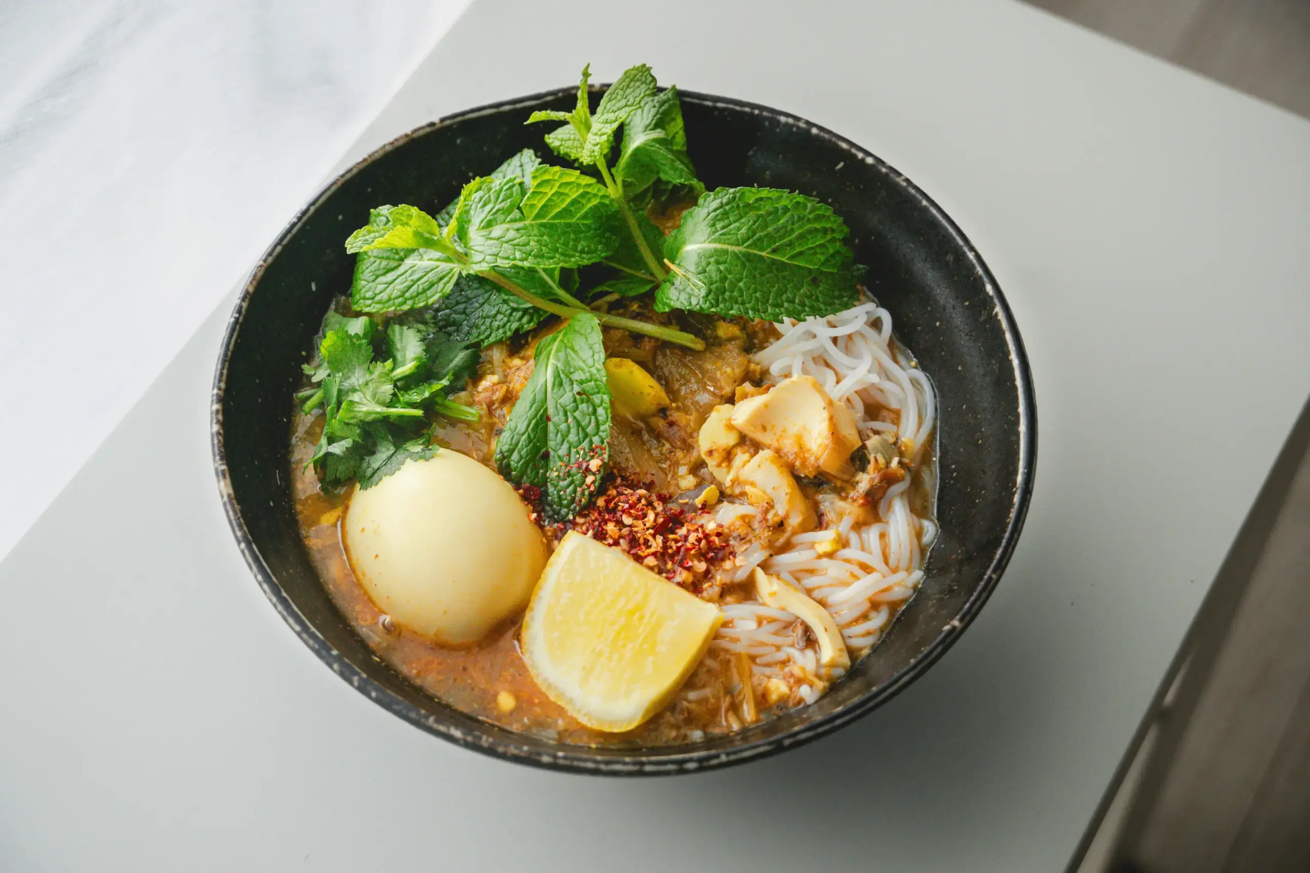 A vibrant bowl of Mohinga features thin rice noodles submerged in a savory fish broth, topped with a hard-boiled egg and fresh mint leaves. The dish is garnished with a bright lemon wedge, chili flakes, and fresh cilantro for a burst of color and flavor.