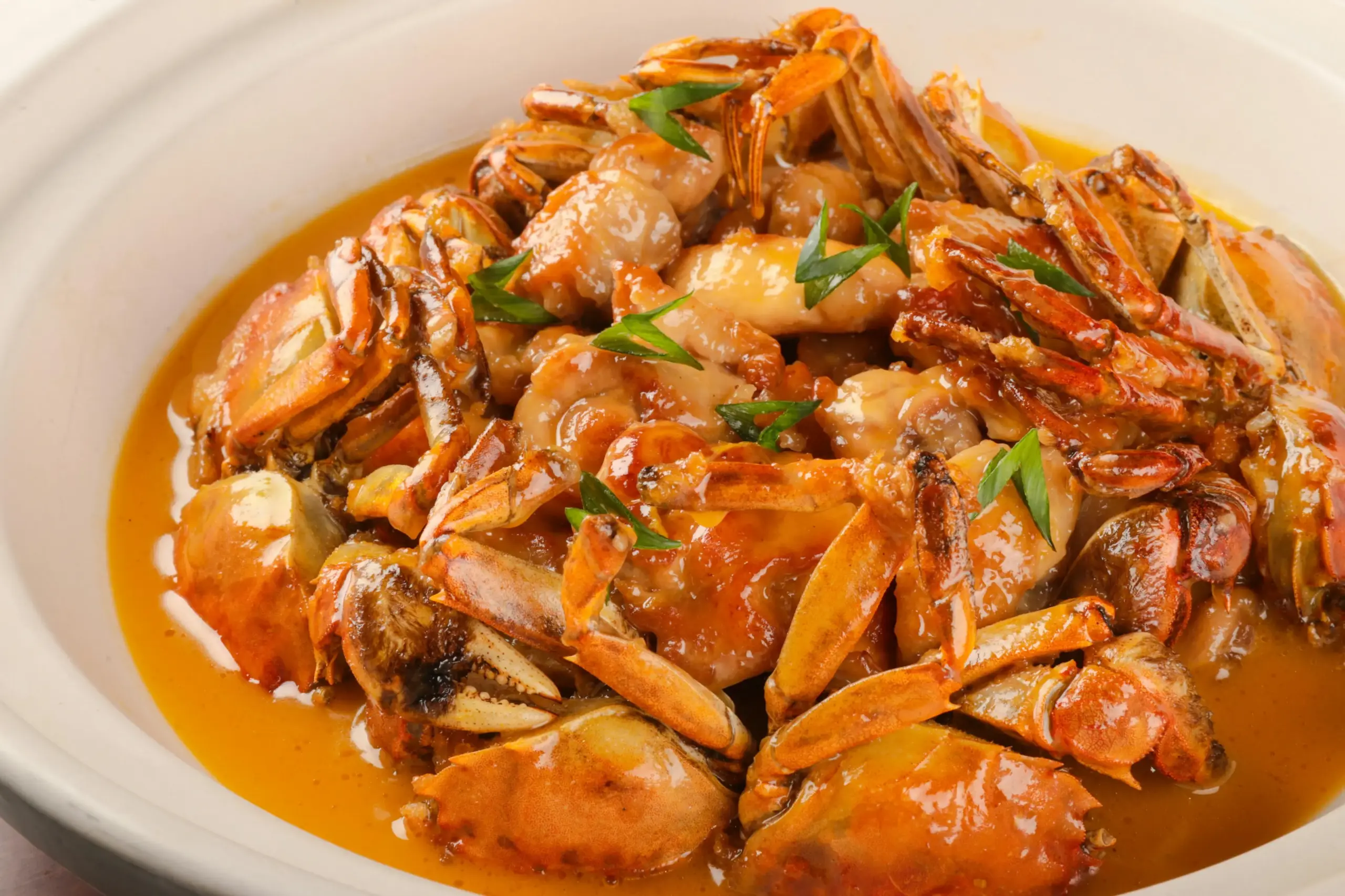 A white ceramic bowl is filled with cooked crab pieces and tender meat simmered in a rich, orange-hued savory sauce. The dish is garnished with sliced green scallions and served in a shallow pool of flavorful broth.