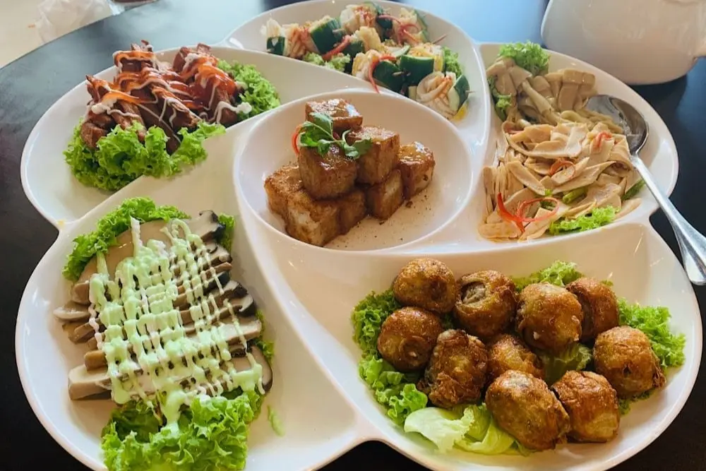 A variety of vegetarian appetizers are arranged on a large, white, flower-shaped platter set on a dark table. The dishes include fried tofu cubes, crispy rolls, sliced mock meats with colorful sauces, and a refreshing cucumber salad.