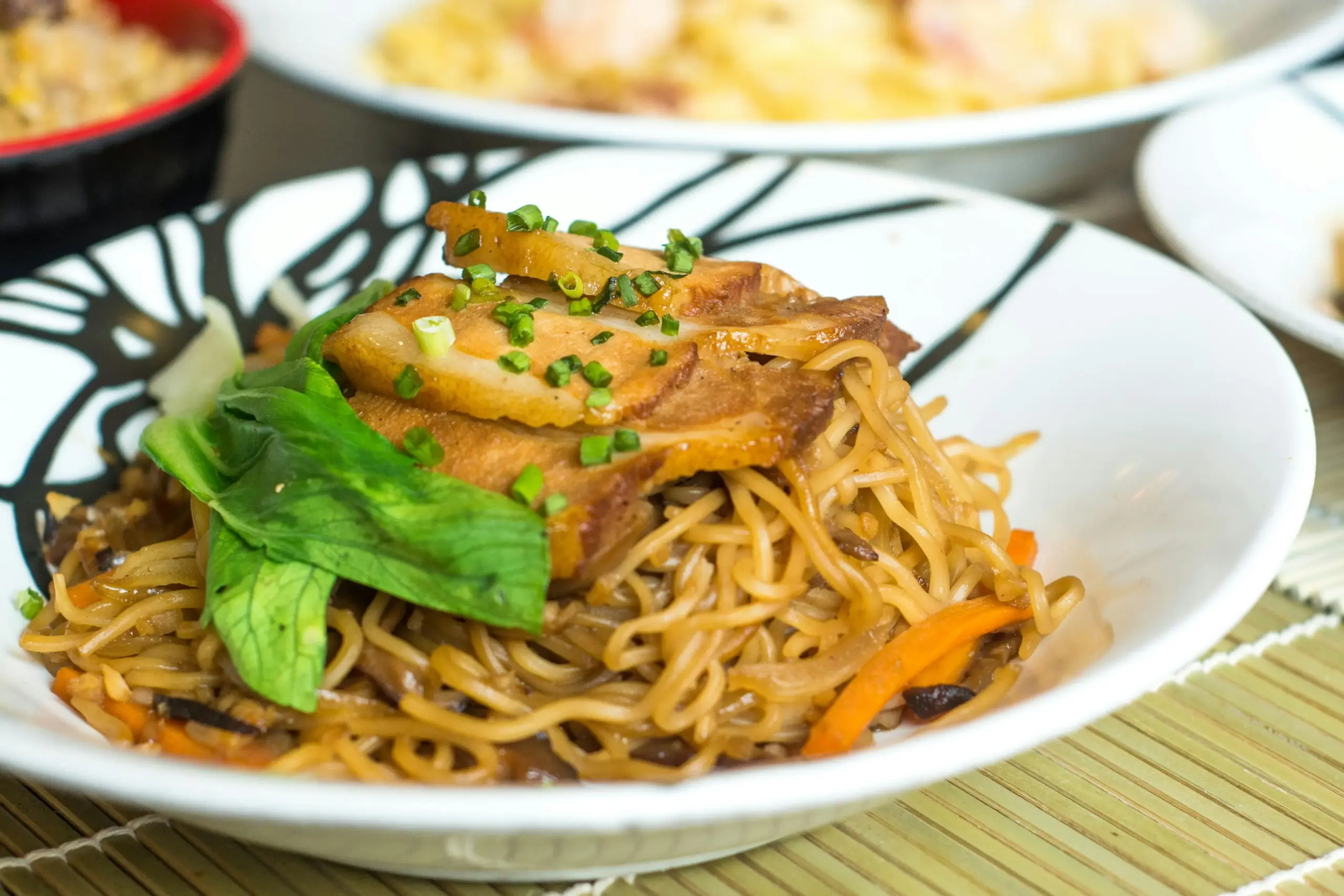 A white bowl holds a mound of stir-fried noodles topped with slices of glazed pork and fresh green vegetables. Other dishes, including fried rice and noodles, are visible in the soft-focus background on a bamboo mat.