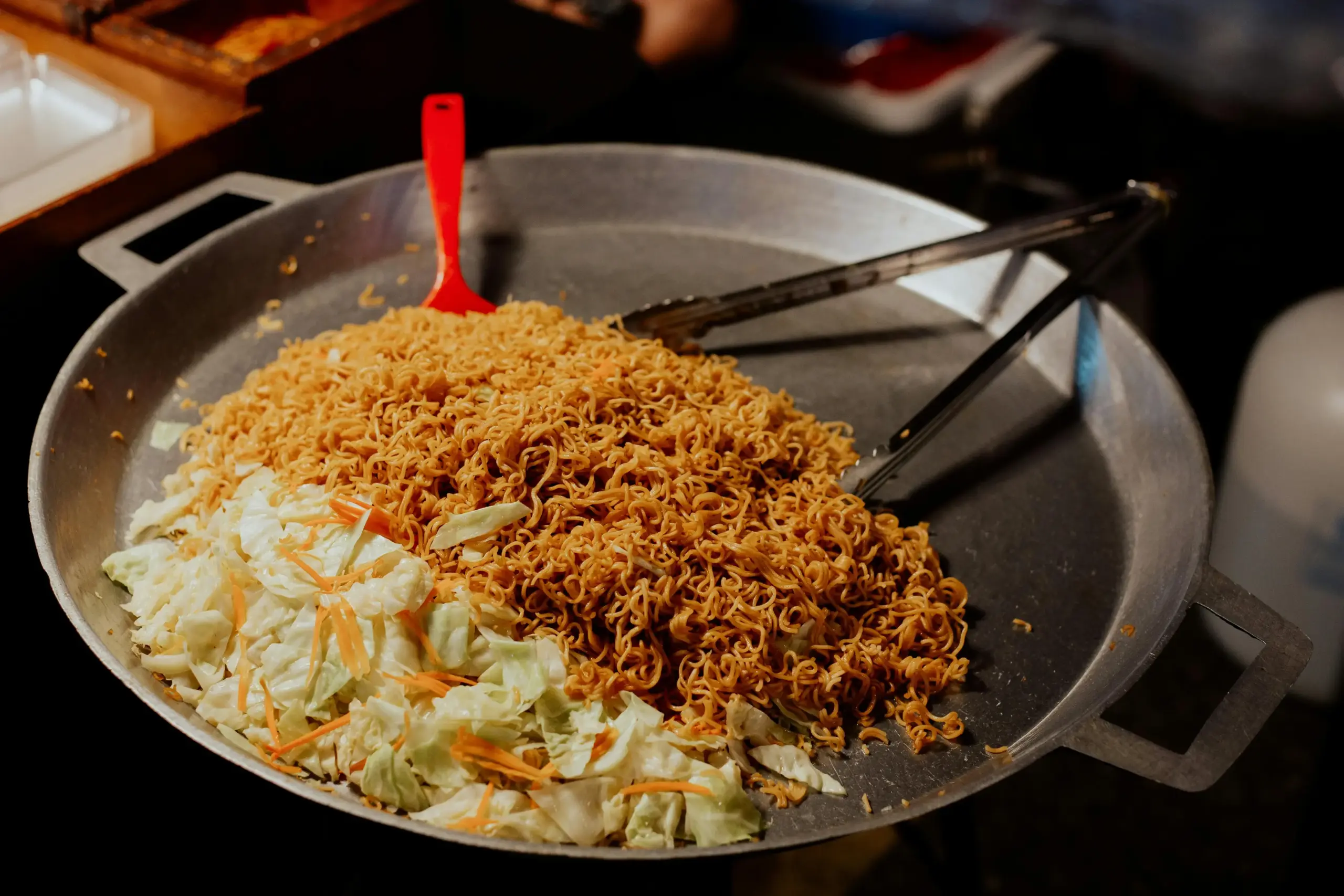 A large metal pan holds a heap of seasoned instant noodles alongside a portion of shredded cabbage and carrots. A red spatula and a pair of metal tongs rest inside the pan, ready for tossing the ingredients together.