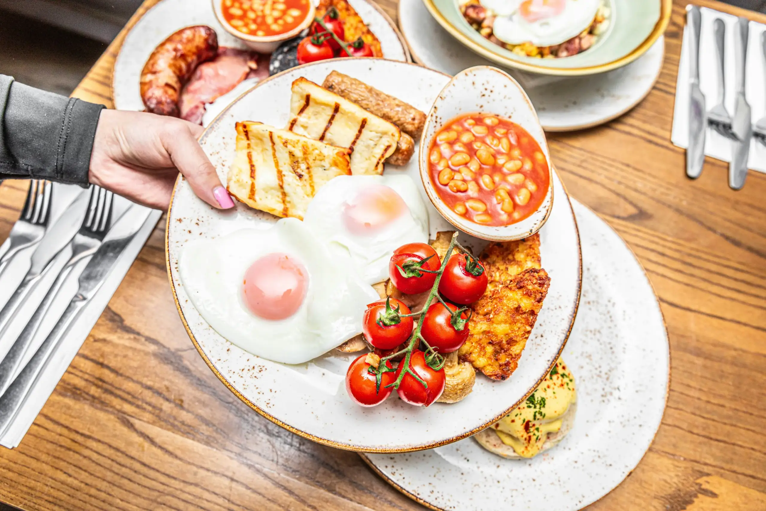 A hand with pink nails holds a plate featuring a hearty breakfast of fried eggs, grilled halloumi, baked beans, and a vine of roasted tomatoes. The wooden table is crowded with additional brunch dishes, including eggs benedict and sausages, alongside sets of silver cutlery.