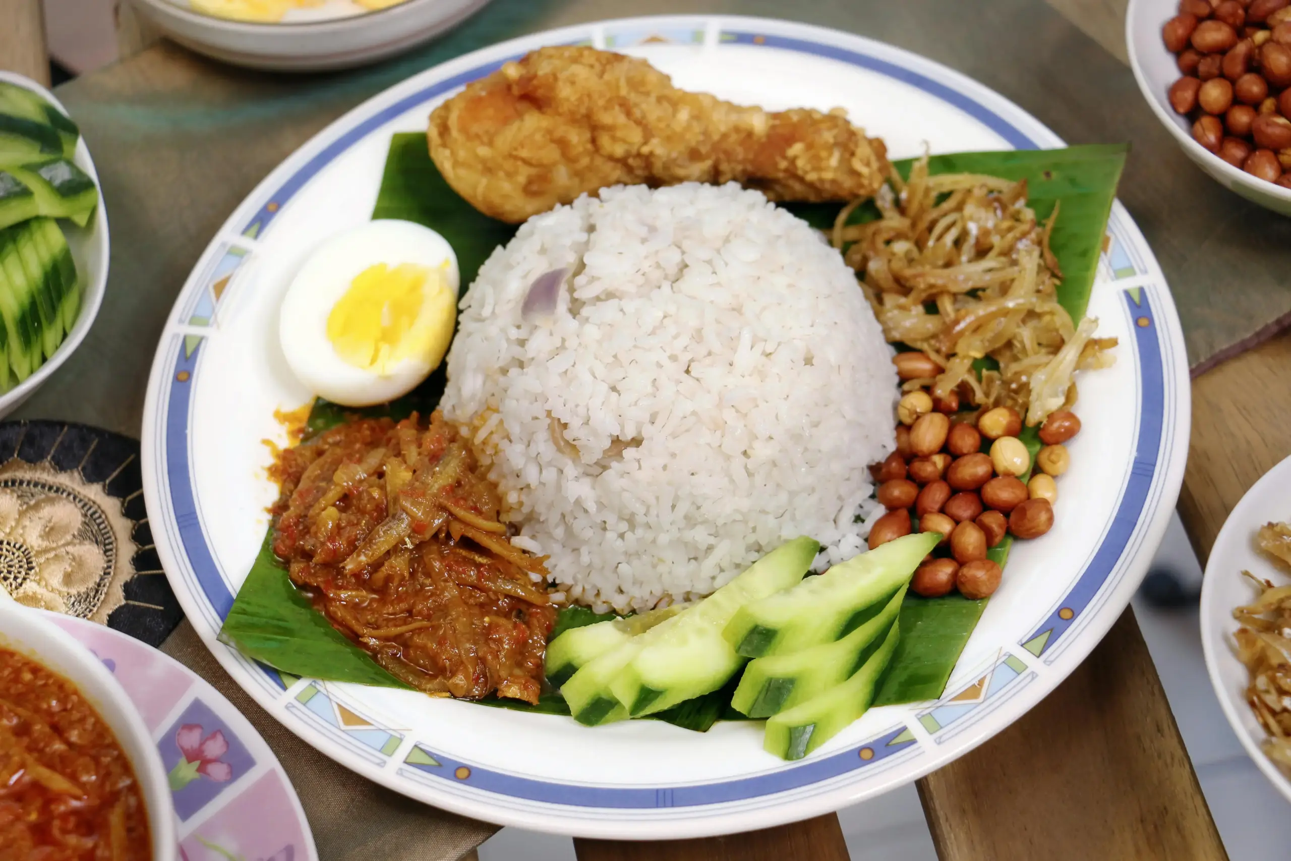 A vibrant plate of Nasi Lemak is presented on a banana leaf, featuring a mound of coconut rice surrounded by fried chicken, sambal, a hard-boiled egg, and classic garnishes like peanuts and cucumber. Several small side dishes containing extra peanuts and spicy sauces accompany the main meal on a wooden tabletop.