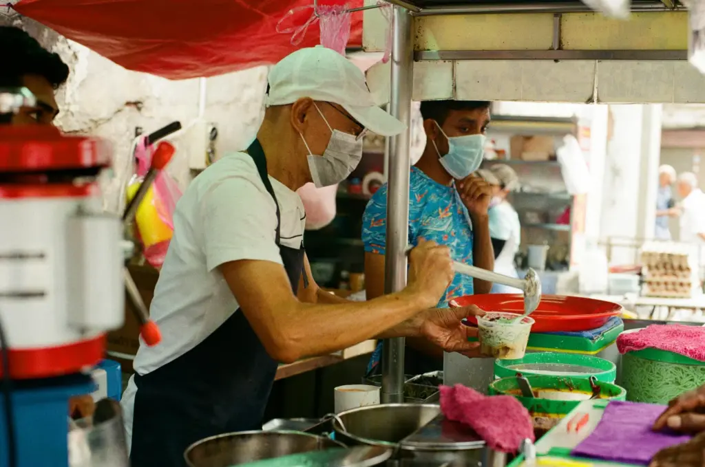 A street food vendor wearing a mask and white cap carefully prepares a colorful dessert in a clear plastic container. The bustling stall features various bowls of ingredients and a red ice shaver in the foreground.