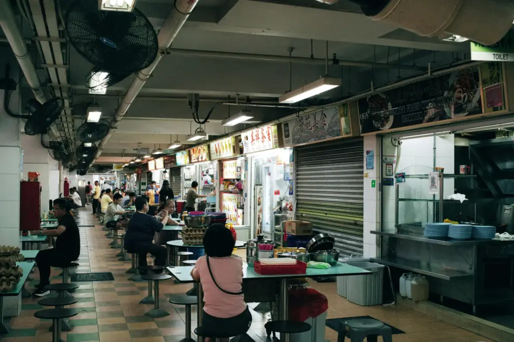 Patrons sit at round tables in a bustling indoor hawker center lined with various food stalls and industrial ceiling fans. The scene captures a slice of daily life, featuring tiled flooring and rows of open-front kitchens serving local cuisine.