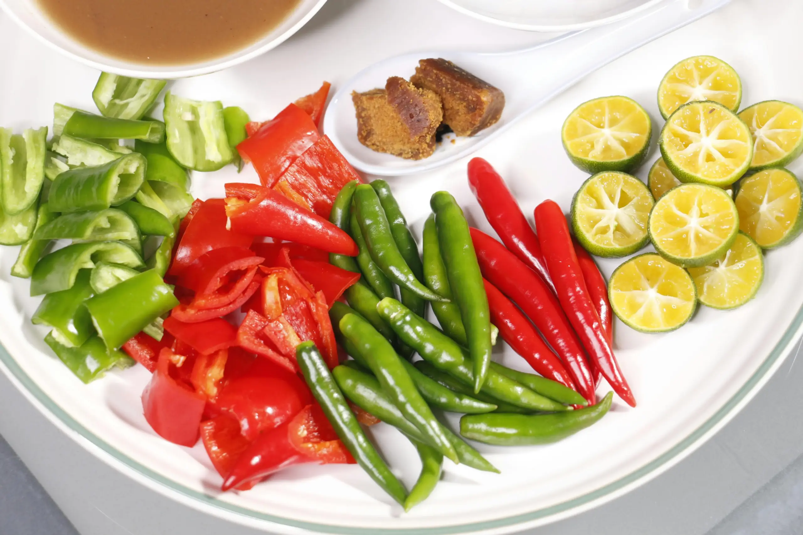 This white plate displays a vibrant assortment of fresh ingredients, including sliced green and red peppers, whole bird's eye chilies, and halved calamansi limes. A small white spoon rests near the top, holding pieces of palm sugar alongside a bowl of dark dipping sauce.
