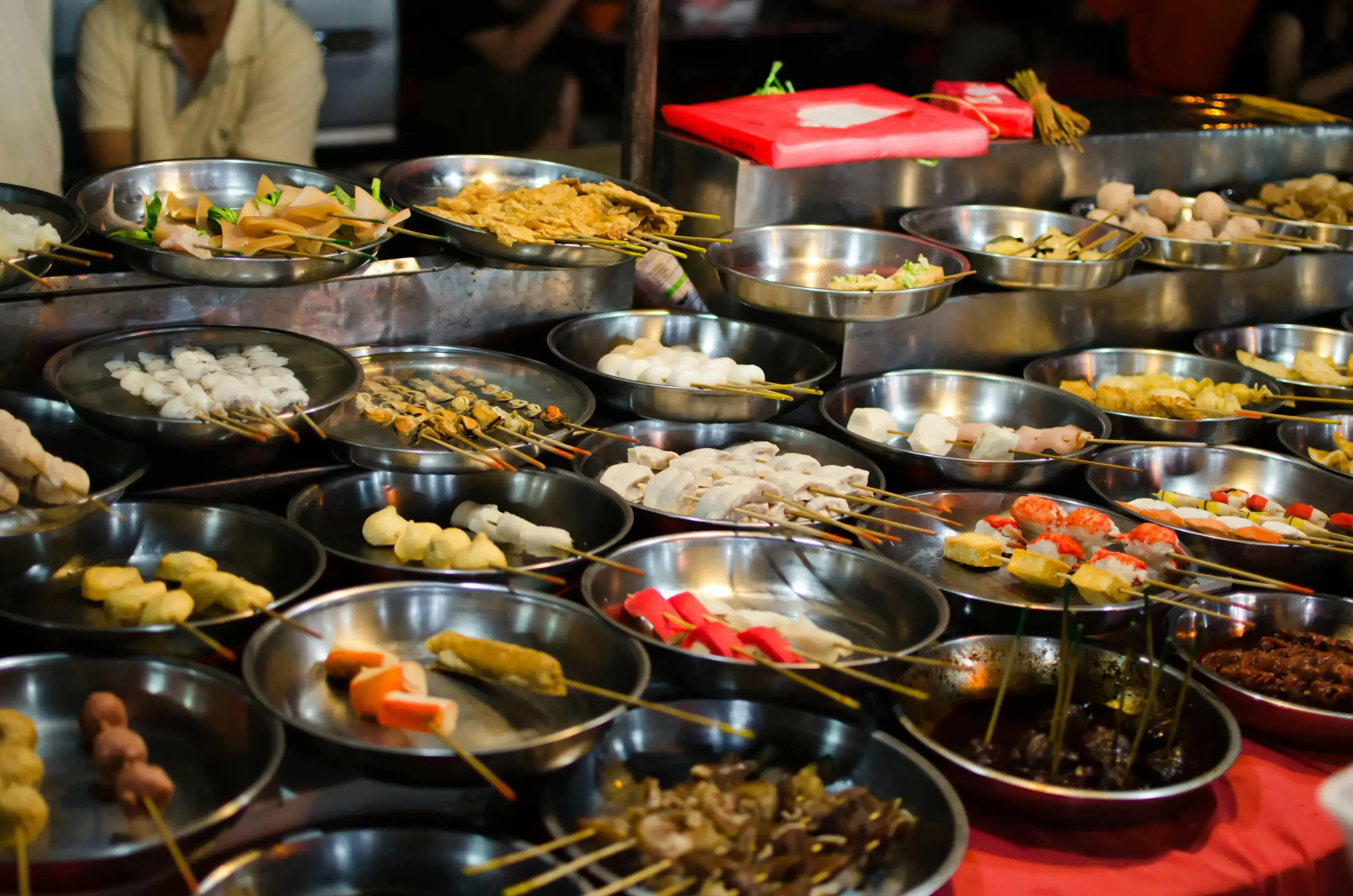 A vibrant street food stall displays a wide variety of skewered ingredients, including fish balls, meats, and seafood, arranged in tiered metal bowls for customers to select. This bustling night market scene captures the diverse and colorful options typical of Southeast Asian outdoor dining culture.