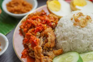 The image shows a vibrant plate of Indonesian fried chicken topped with a generous layer of spicy red sambal. Accompanying the chicken is a mound of white rice sprinkled with fried shallots, halved hard-boiled eggs, and fresh cucumber slices.