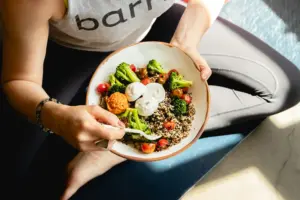 In the image, a person in athletic wear sits cross-legged while holding a vibrant grain bowl. The dish is packed with quinoa, broccoli, cherry tomatoes, and topped with dollops of creamy cheese or sauce.