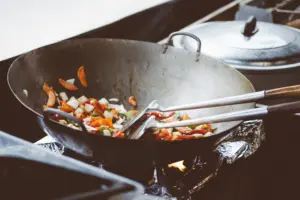 A large, well-used metal wok sits over an open flame, filled with a colorful mix of chopped vegetables like carrots, onions, and peppers. Two long-handled metal spatulas rest inside the pan, ready to stir the ingredients as they sizzle over the high heat.