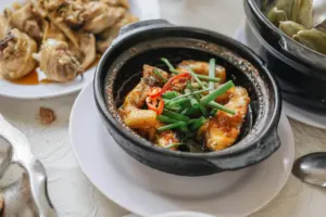 A black clay pot sits at the center of the frame, filled with pieces of glazed fish topped with fresh green scallions and sliced red chilies. Several other dishes, including a plate of braised chicken, are spread across the white textured tablecloth in a bright, inviting dining setting.
