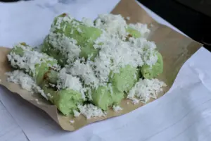 The image shows several cylindrical, light-green rice cakes generously topped with shredded white coconut. These traditional snacks are served on a piece of brown paper resting atop a white surface with printed text.