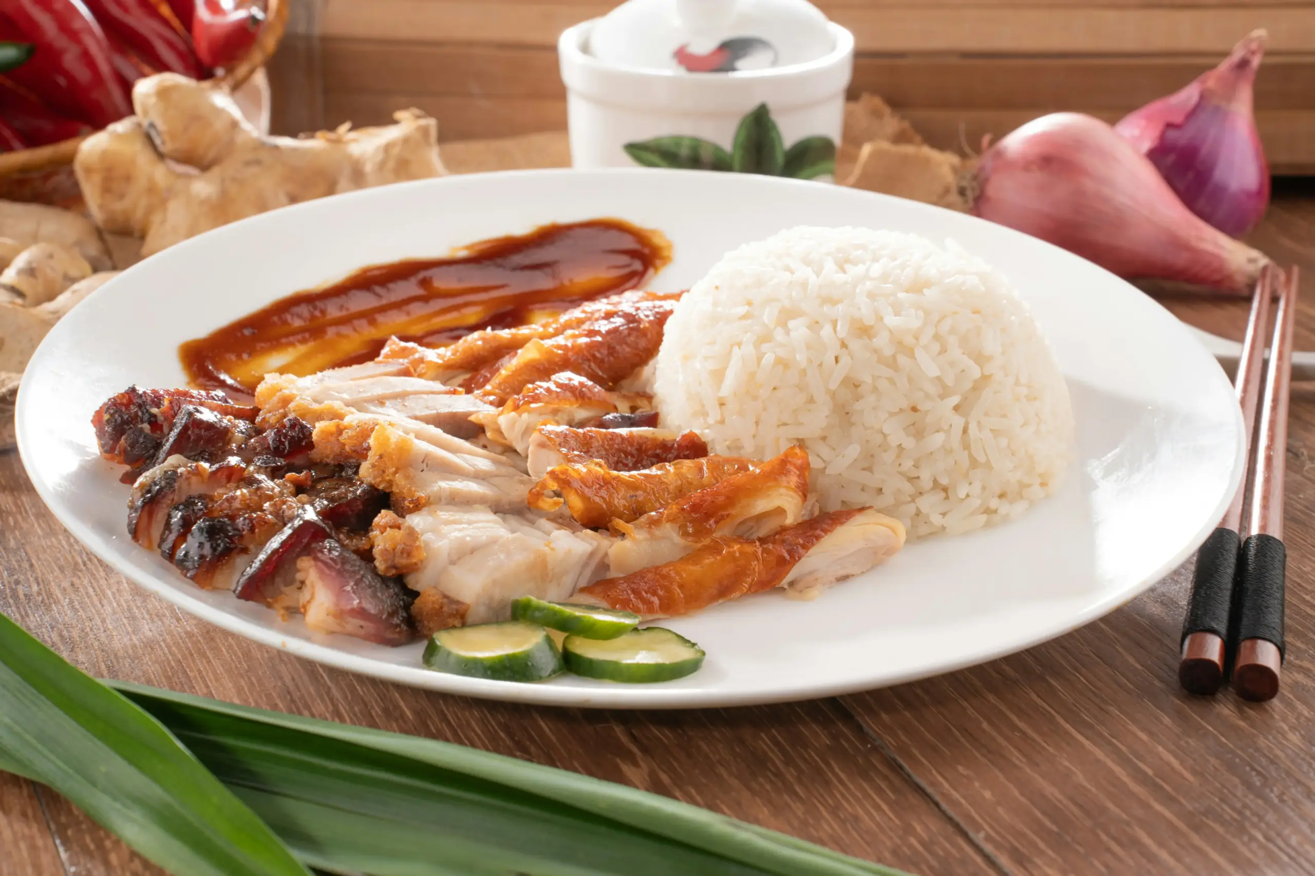The image depicts a traditional Singaporean dish featuring sliced roasted meats, a mound of white rice, and cucumber garnishes on a white plate. In the background, fresh ingredients like ginger, shallots, and pandan leaves are artfully arranged on a wooden surface alongside a pair of chopsticks and a small ceramic bowl.