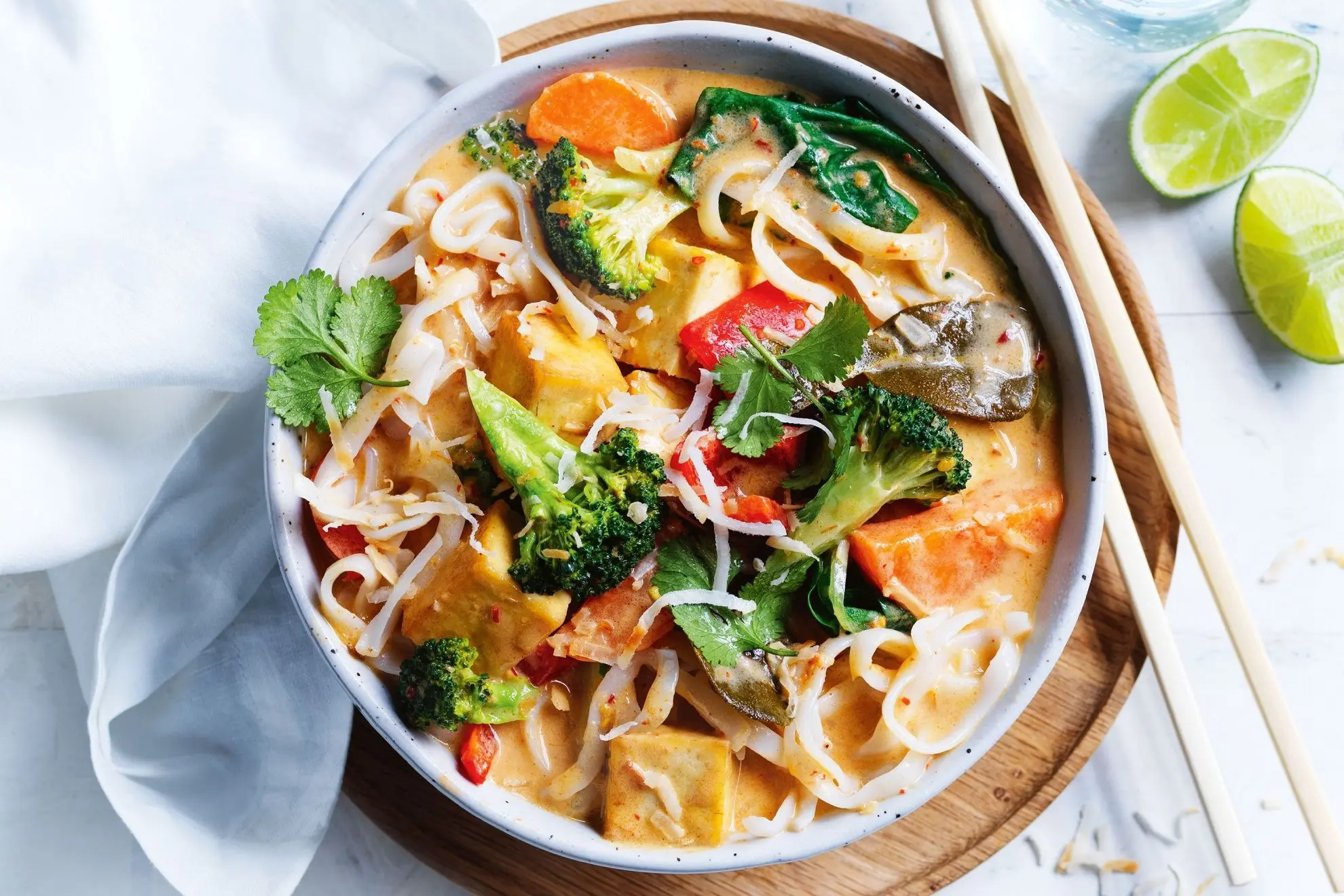 The image shows a vibrant bowl of laksa filled with rice noodles, tofu cubes, broccoli, and red peppers in a creamy coconut broth. It is served on a wooden board with chopsticks and fresh lime wedges on the side.