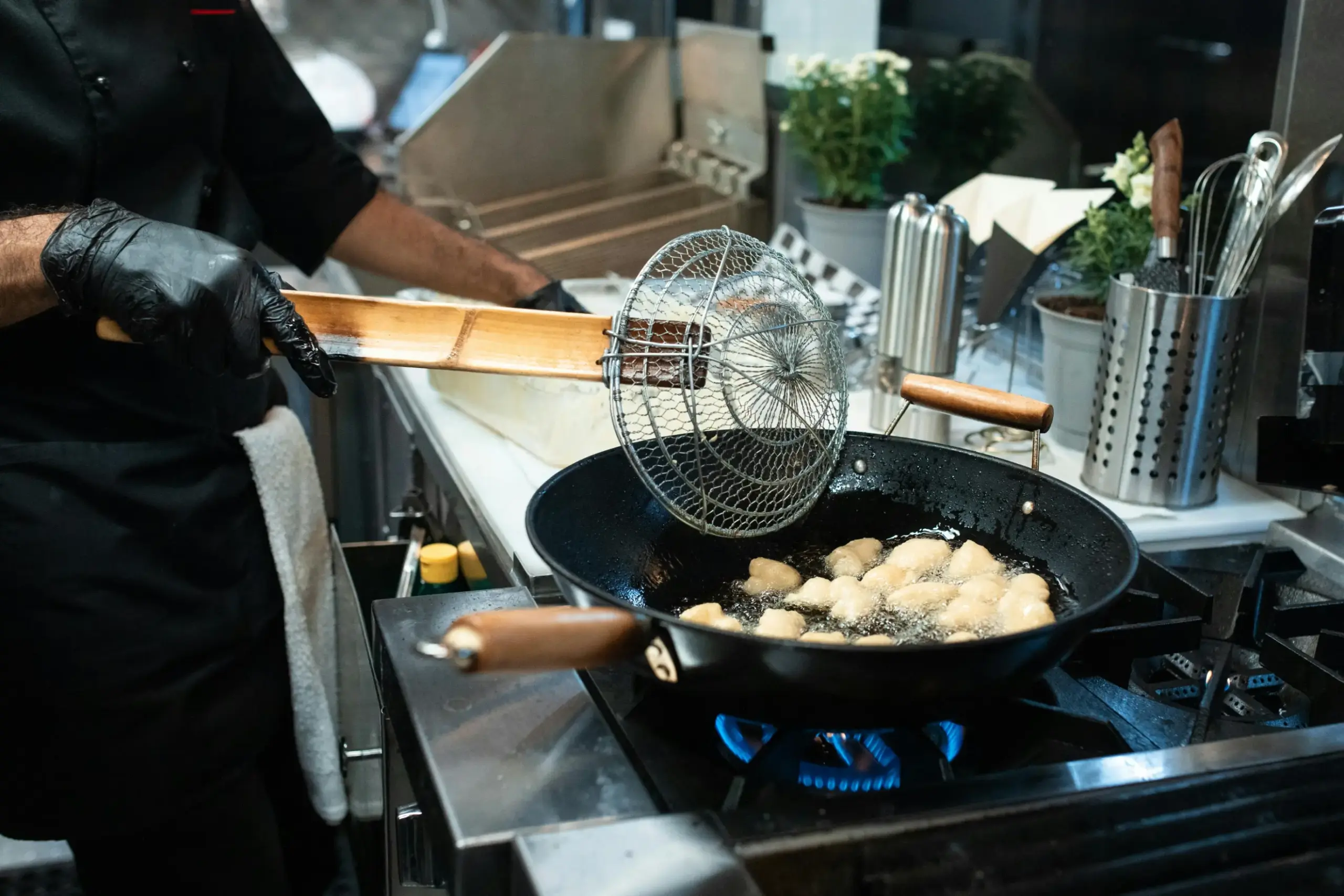 A chef wearing black gloves uses a bamboo-handled spider skimmer to lift golden, fried dough pieces from a bubbling wok. The scene is set in a professional kitchen over a bright blue gas flame, surrounded by stainless steel tools and potted herbs.