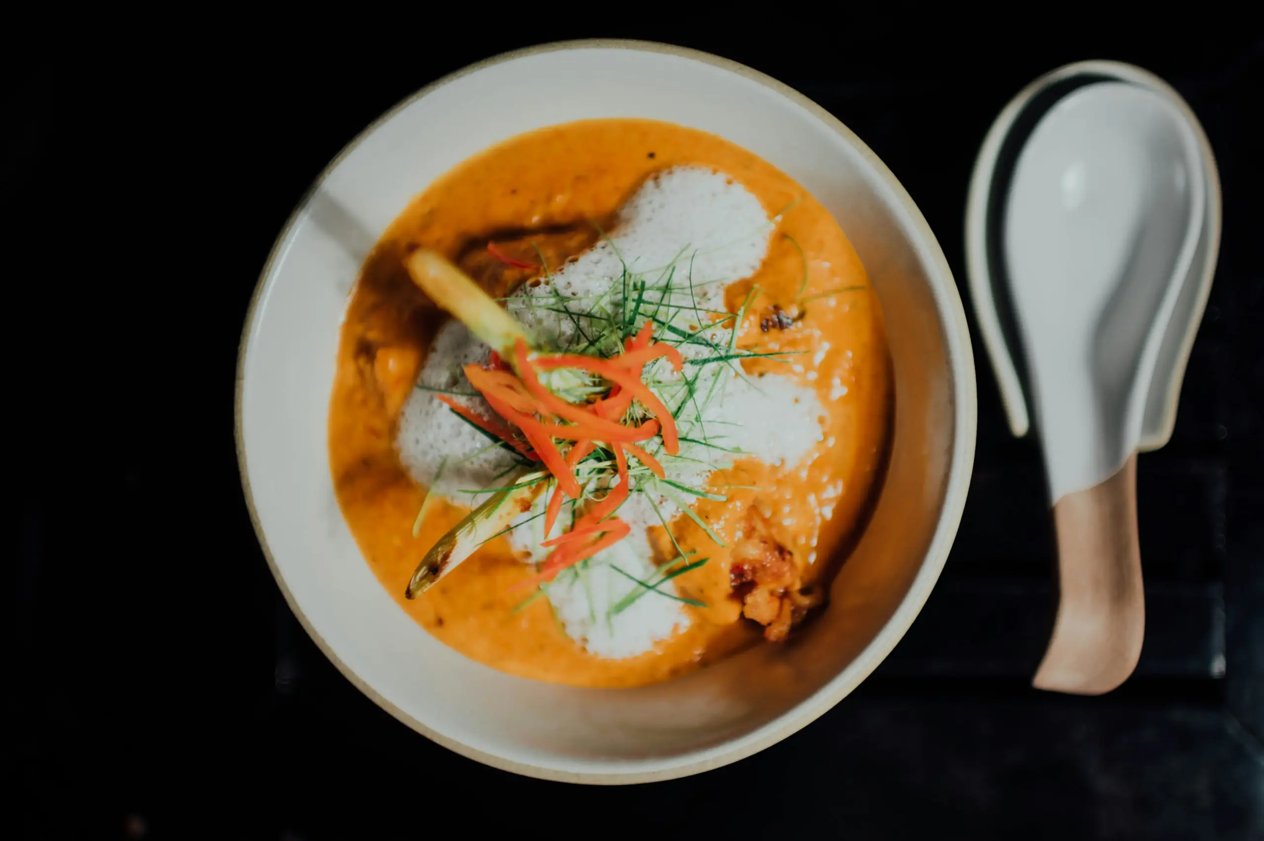 An overhead shot features a bowl of rich, orange laksa topped with white foam, thin green herbs, and bright red chili slivers. Beside the bowl sits a modern white and wood-toned spoon against a dark, minimalist background.