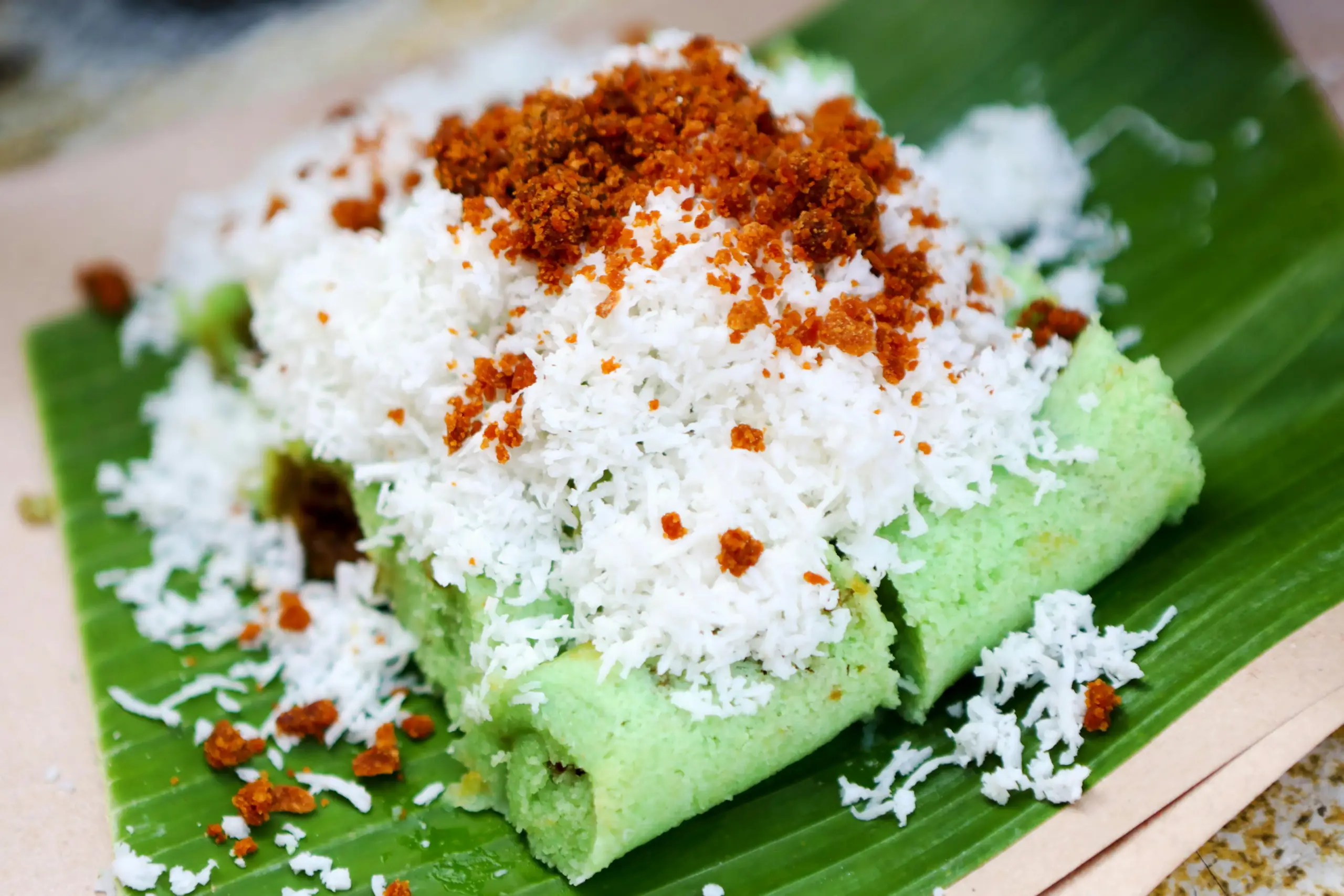 The image features a vibrant green putu piring dessert served on a fresh banana leaf. The steamed rice cakes are generously topped with a mountain of shredded coconut and crumbles of palm sugar.