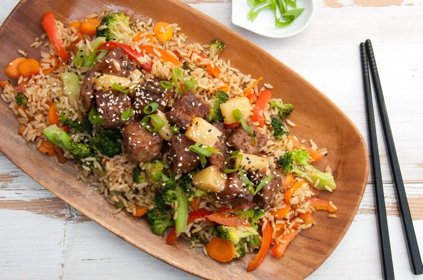 The image features a vibrant dish of fried rice topped with breaded mock chicken, pineapple chunks, broccoli, carrots, and red bell peppers. This colorful meal is served on an oval wooden platter and garnished with sesame seeds and green onions, accompanied by black chopsticks on the side.