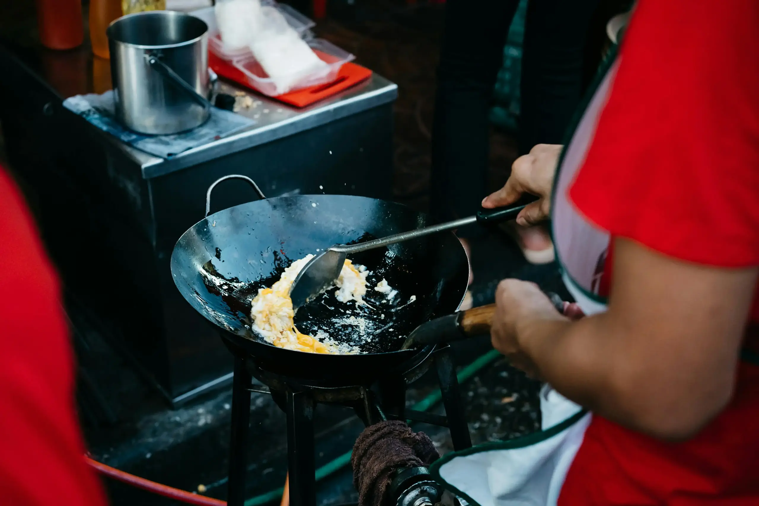 A cook in a red shirt uses a metal spatula to stir-fry eggs in a large, black wok over a high-heat burner. The close-up shot captures the busy atmosphere of an outdoor food stall, with stainless steel equipment and ingredients visible in the background.