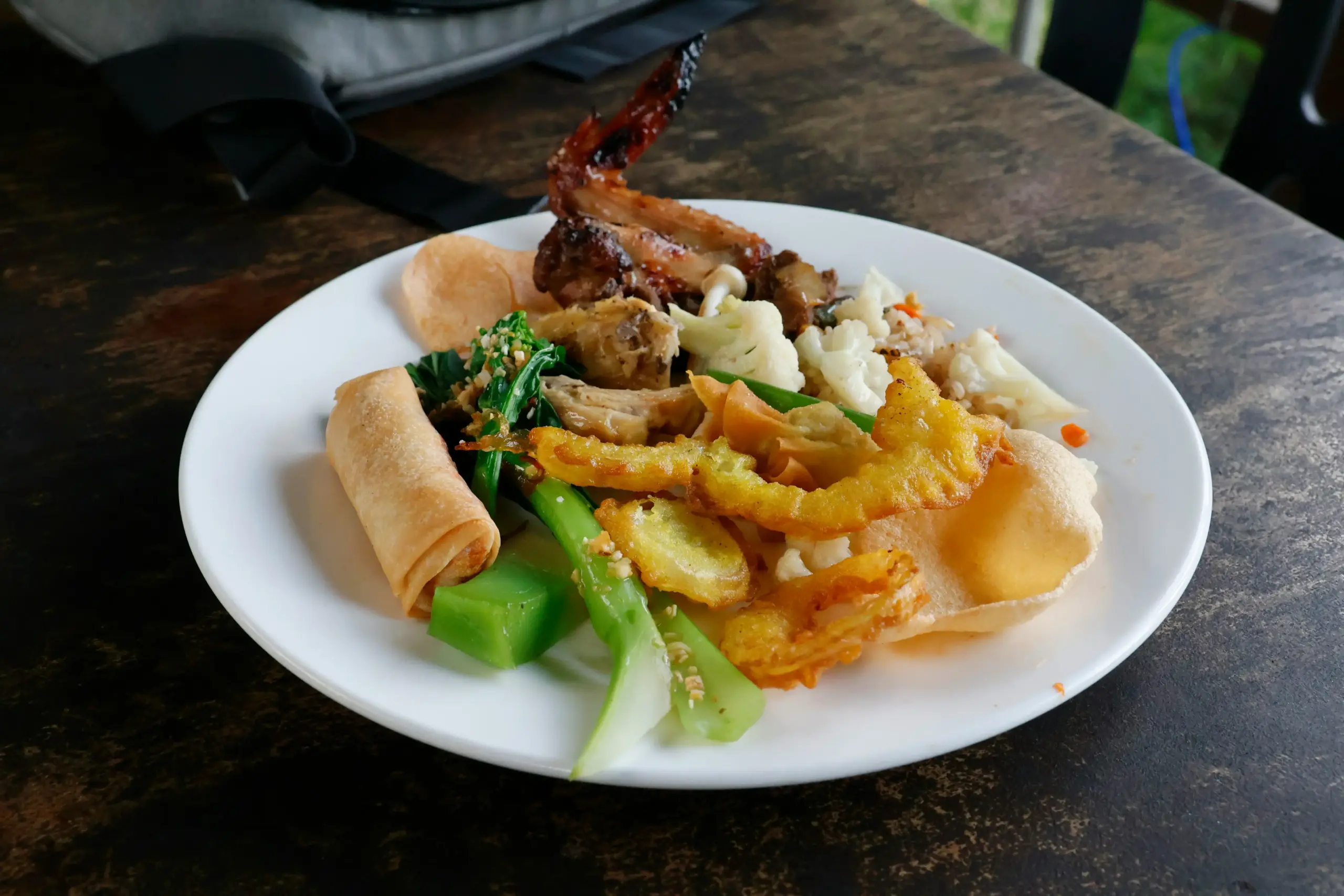 A white plate holds a colorful variety of dishes, including a crispy spring roll, grilled chicken wing, and sautéed green vegetables. Scattered among the meal are golden fried snacks and florets of cauliflower, all served on a dark, rustic wooden table.