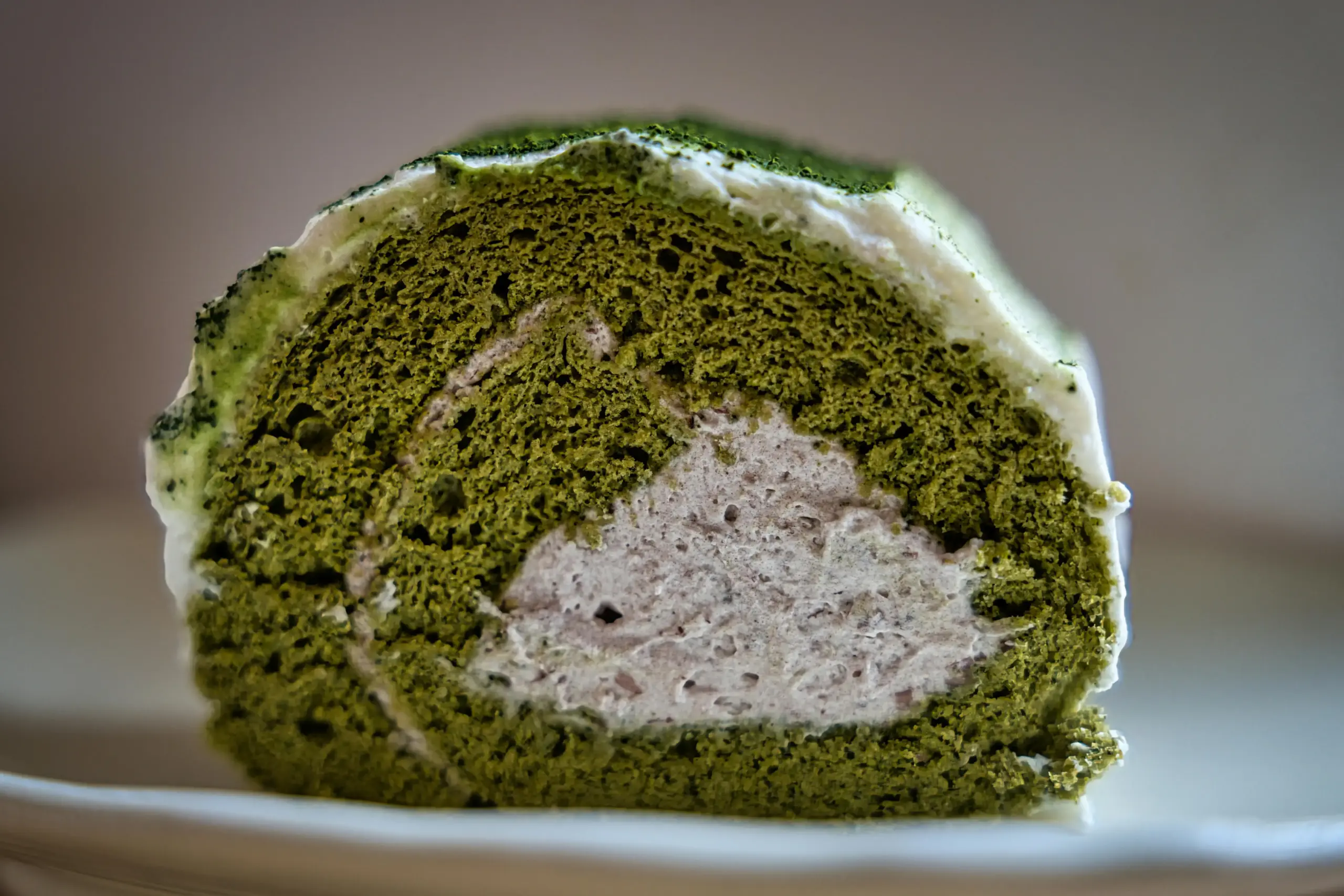 The image shows a vibrant green matcha swiss roll is filled with a light, creamy center and dusted with fine green tea powder. The sponge cake features a porous, aerated texture and is coated in a thin layer of white icing.