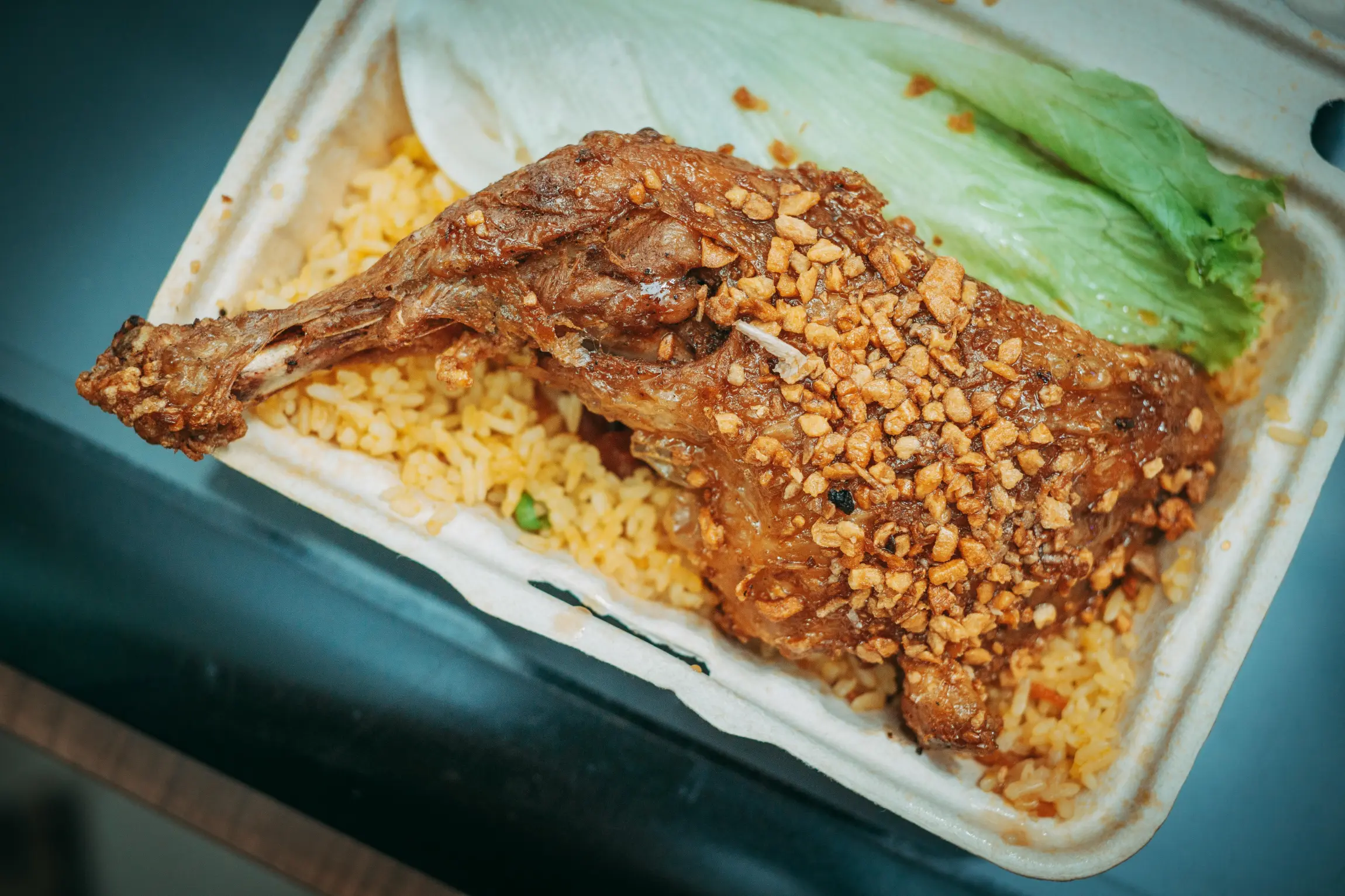 The image displays a take-out container filled with a hearty meal of seasoned rice and a large, roasted duck leg topped with crispy garlic. A fresh green lettuce leaf rests on the side, providing a bright contrast to the savory, golden-brown tones of the dish.