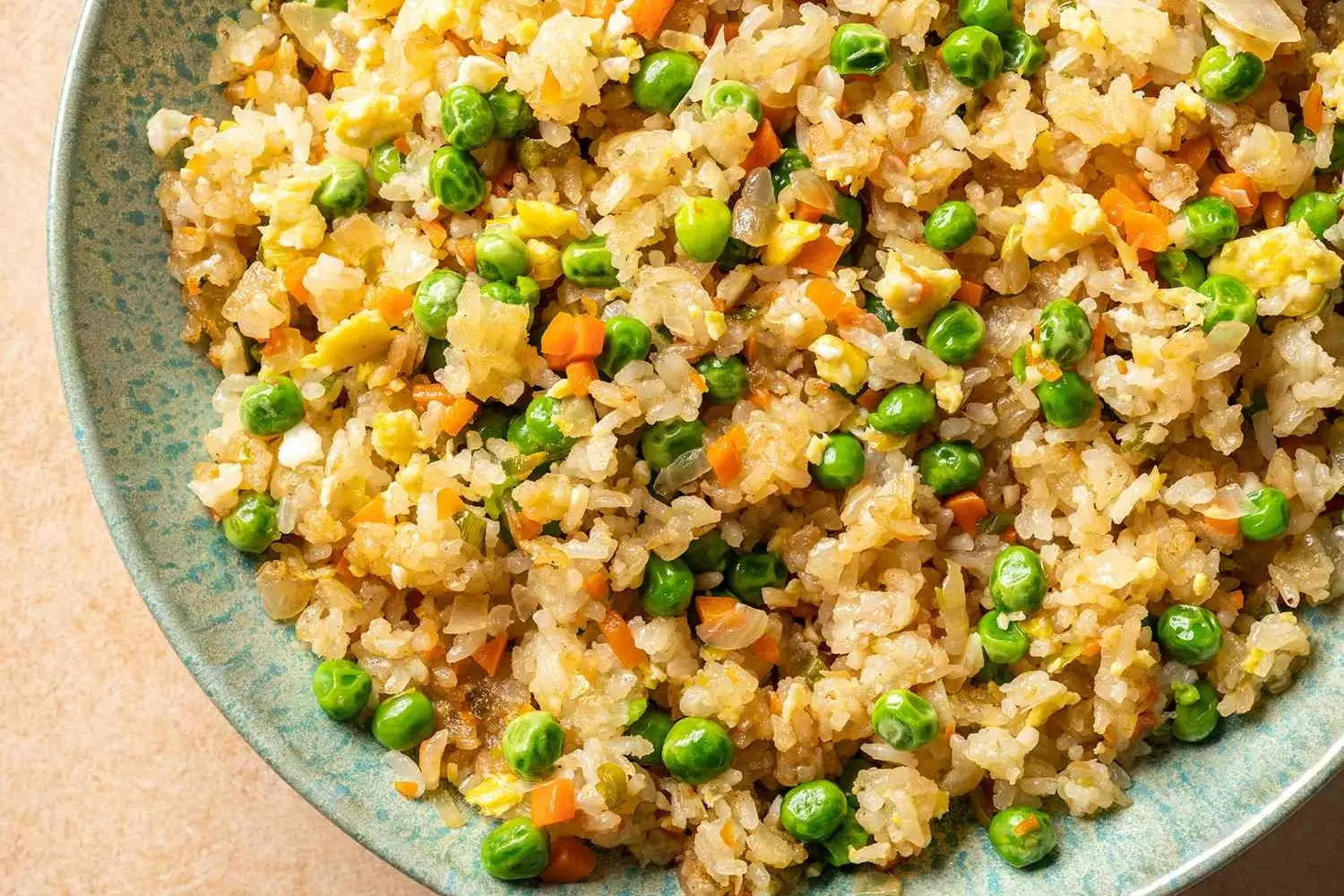 The image shows a top-down view of a textured teal bowl filled with vibrant vegetable fried rice. The dish is a colorful mix of golden rice grains, bright green peas, diced orange carrots, and bits of scrambled egg.