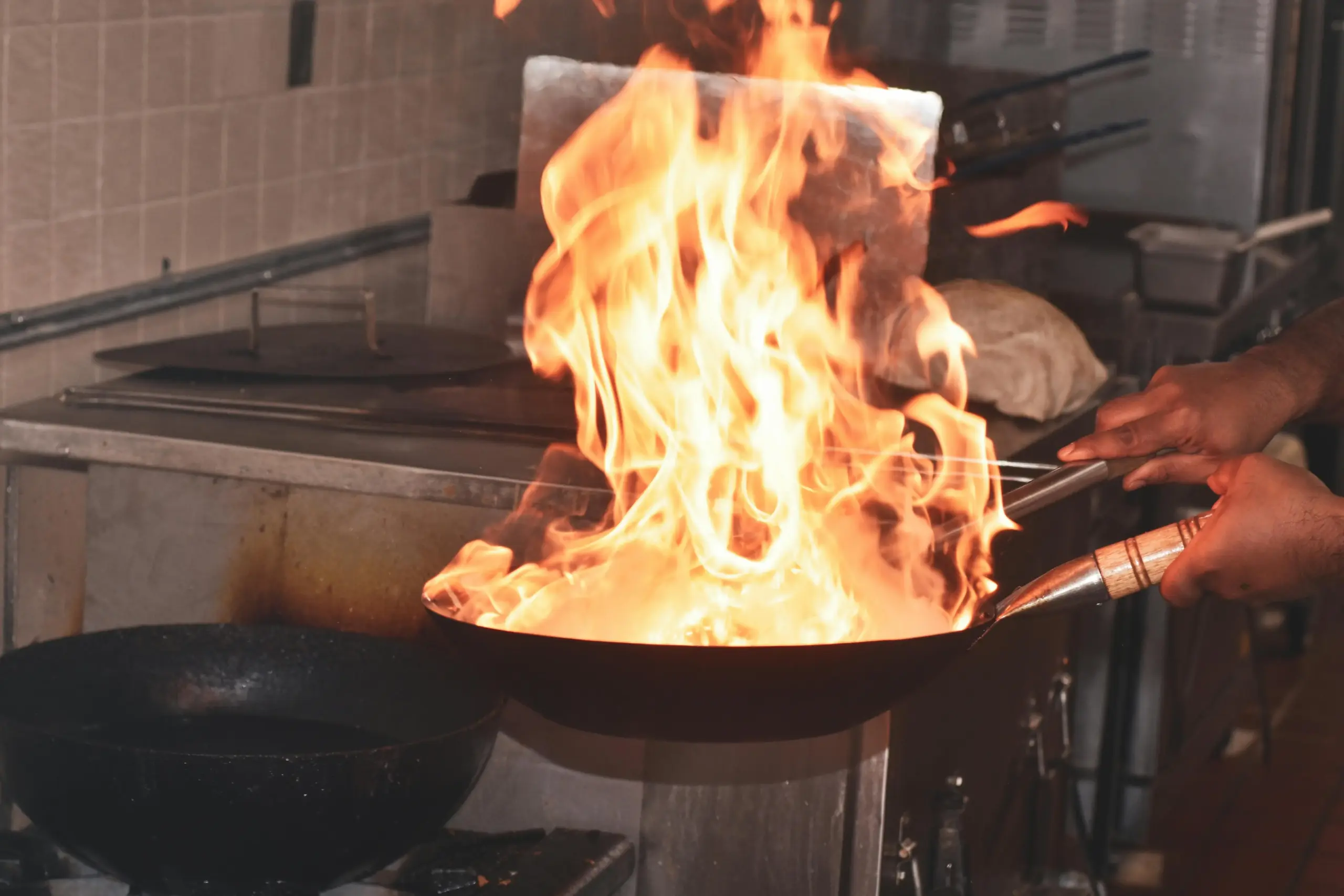 A chef skillfully tosses a wok in a professional kitchen, sending a vibrant burst of orange flames high into the air. This high-heat cooking technique creates a dramatic display of fire and motion against the industrial stainless steel background.