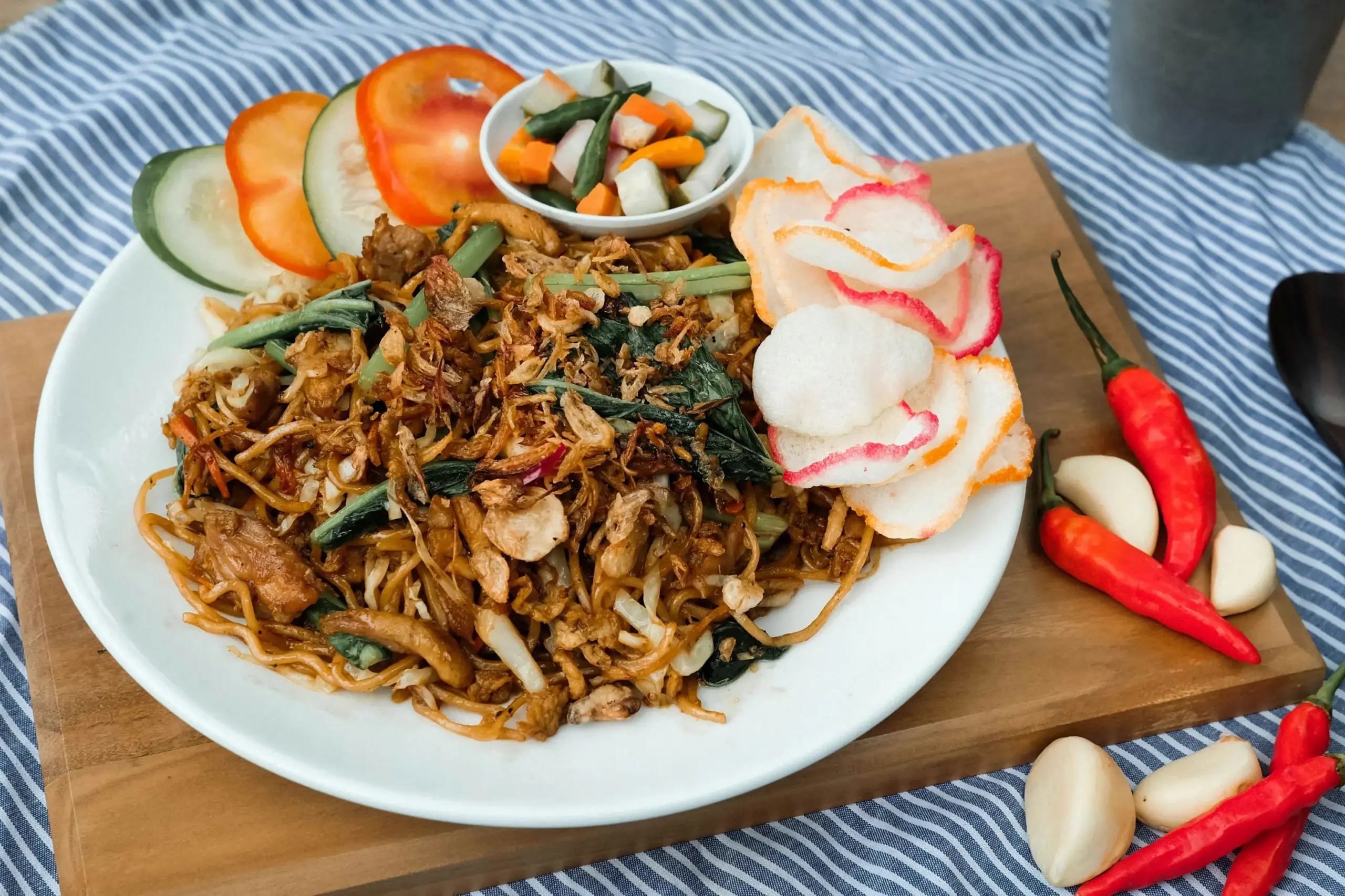 A plate of stir-fried noodles topped with crispy fried shallots is served with shrimp crackers, fresh cucumber, and tomato slices. Beside the plate, whole garlic cloves and vibrant red chili peppers rest on a wooden board.