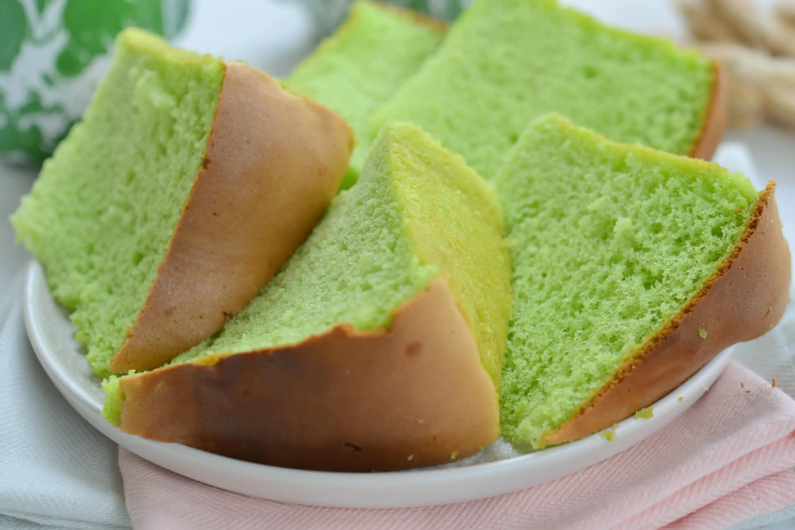 The image features several light green, fluffy slices of cake with a golden-brown crust arranged on a white plate. The slices are set against a soft background with a pink napkin and a hint of a green pattern, emphasizing their airy, sponge-like texture.