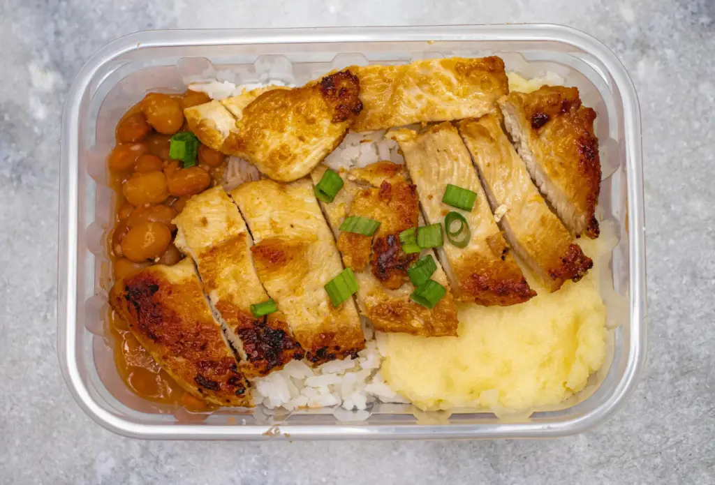 The image shows a clear plastic container filled with sliced grilled chicken, white rice, beans, and mashed potatoes. Sprinkled with chopped green onions, the meal is presented from a top-down perspective on a textured gray surface.