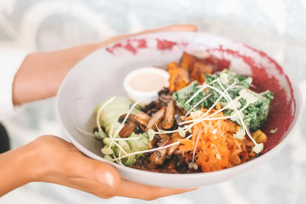 Two hands hold a large, shallow bowl filled with a colorful nourishing bowl in the meal features sautéed mushrooms, sliced avocado, shredded carrots, and leafy greens topped with delicate sprouts and a side of creamy dressing.