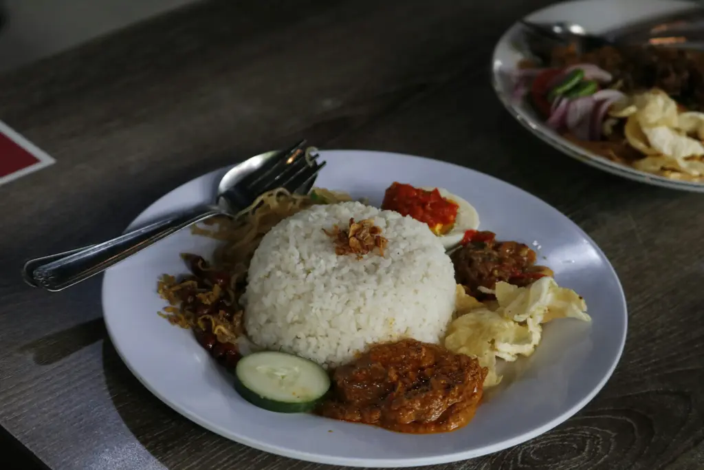 A mound of white rice topped with fried shallots serves as the centerpiece of this Nasi Lemak plate, which is accompanied by several traditional sides like beef rendang and sambal. Surrounding the rice are textures of crunchy peanuts, anchovies, and crackers, rounded out by a fresh cucumber slice and a hard-boiled egg.