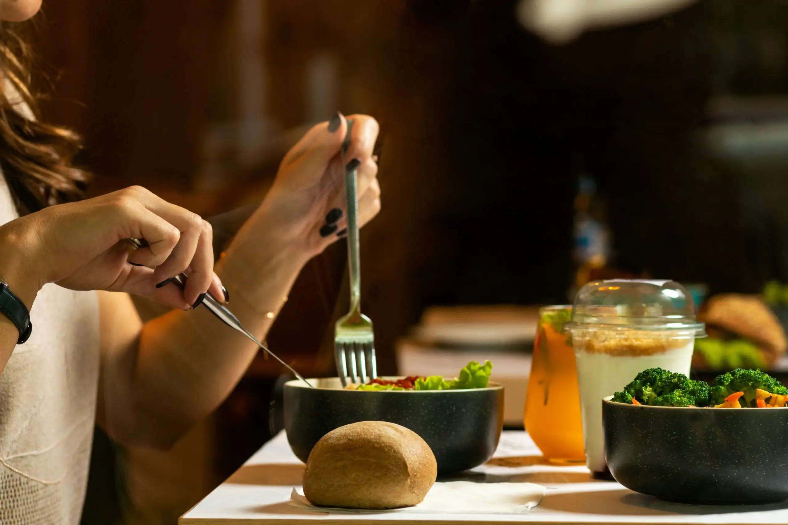 In the image, a person is shown using a fork and knife to eat a fresh salad from a dark bowl. The table is also set with a bread roll, a side of broccoli, and two refreshing drinks in a warm, dimly lit setting.