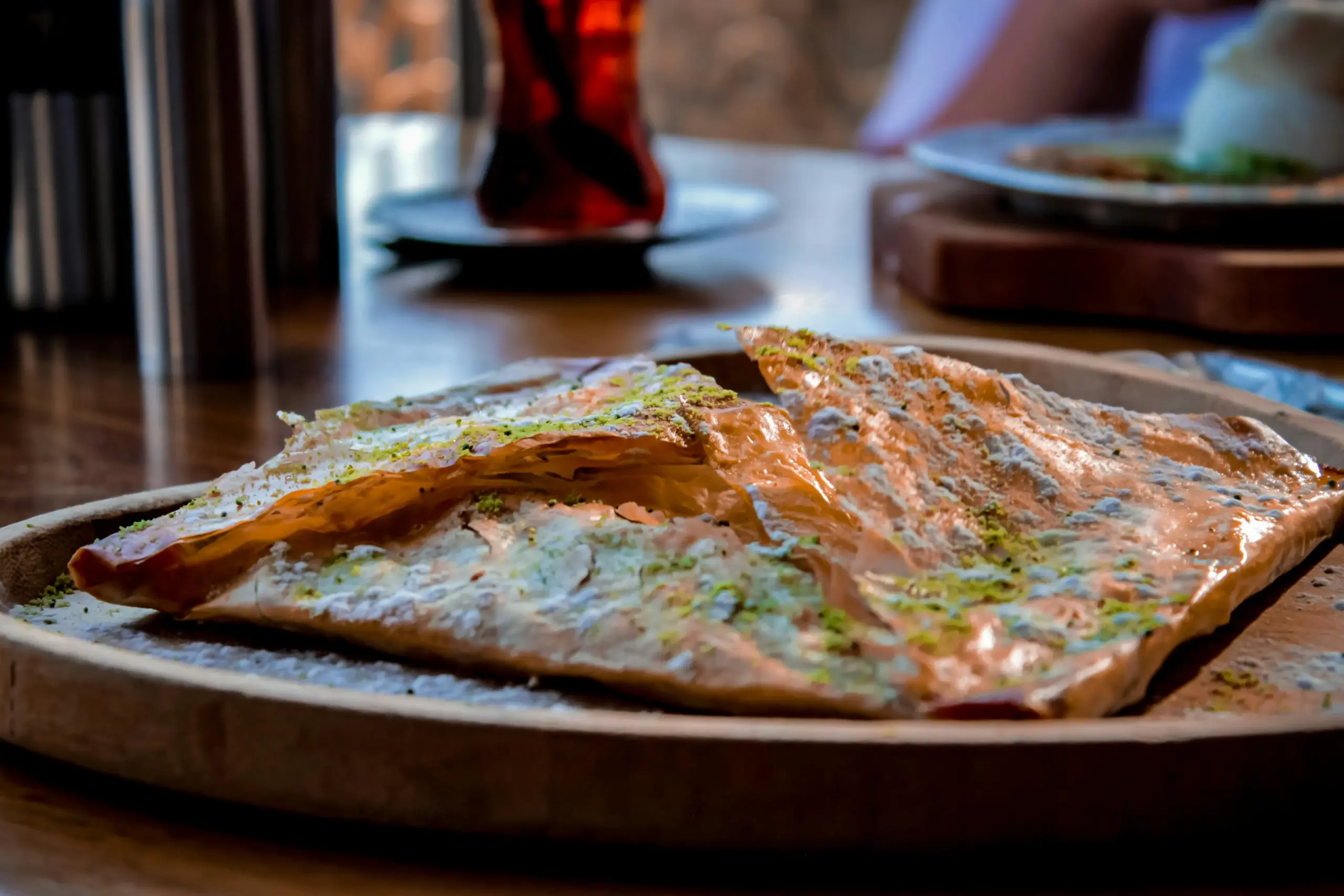 A golden-brown, crispy pastry dusted with bright green pistachio crumbles sits on a rustic wooden tray. In the blurred background, a glass of dark tea and another plated dish suggest a warm, inviting cafe setting.